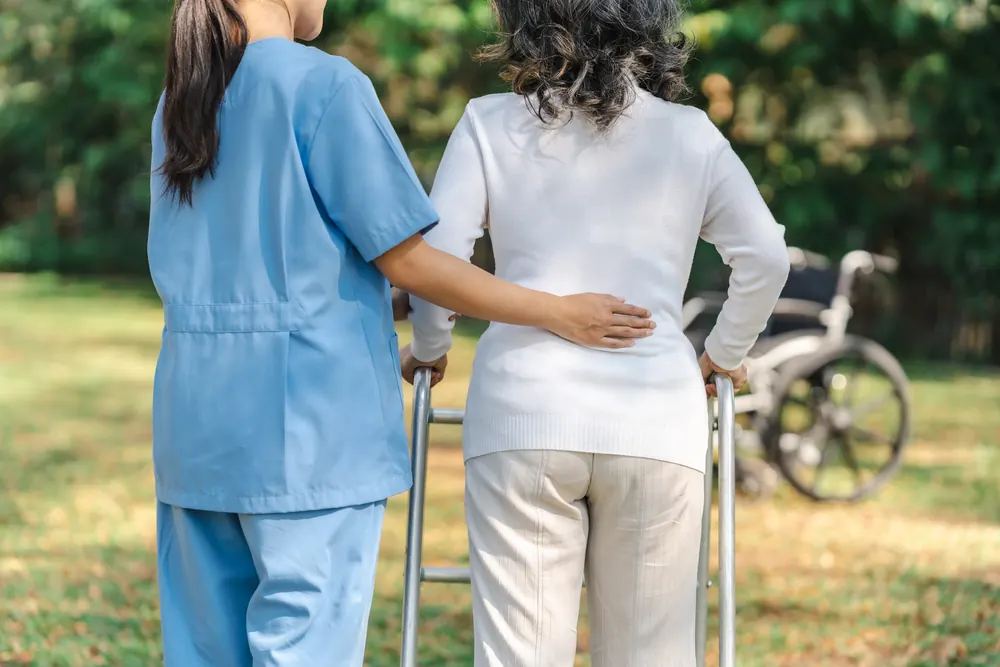 Pflegerin begleitet eine Seniorin mit Rollator im Park, von hinten